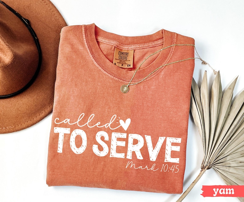 Pu&ograve; includere: Una maglietta Comfort Colors color igname con le parole "called TO SERVE" in bianco, insieme al versetto biblico Marco 10:45. Una collana d'oro e un cappello marrone sono anche nell'immagine.