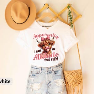 May include: White t-shirt with a cartoon Highland cow wearing a pink bow and the text "Apparently I have an attitude who knew".