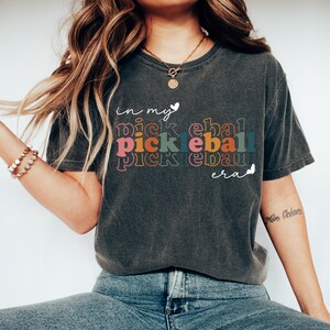 Może przedstawiać: Szary t-shirt z nadrukiem "in my pickleball era" w tęczowych kolorach. Tekst jest ułożony pionowo, a słowo "pickleball" powtarza się trzy razy.