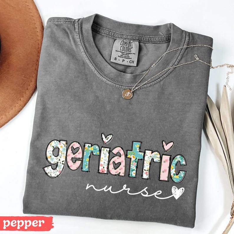 Pode incluir: Uma camiseta cinza Comfort Colors com as palavras "geriatric nurse" em um design floral. A camisa tem gola redonda e mangas curtas. Um colar de prata com um pequeno pingente est&aacute; drapeado sobre a camisa.