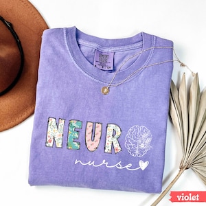 Puede incluir: Una camiseta violeta Comfort Colors con las palabras "NEURO nurse" en un dise&ntilde;o floral. La palabra "NEURO" est&aacute; en un estilo de letras en bloque con un patr&oacute;n floral. Un gr&aacute;fico de cerebro blanco con flores est&aacute; en el lado derecho de la camisa.