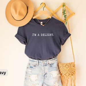 May include: A navy blue t-shirt with the text "I'M A DELIGHT." printed in white. The shirt is hanging on a wooden rack with a straw bag and a hat.