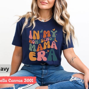 May include: A navy blue t-shirt with the text "In My Homeschool Mama Era" in colorful, wavy letters. The shirt is being worn by a person with long blonde hair.