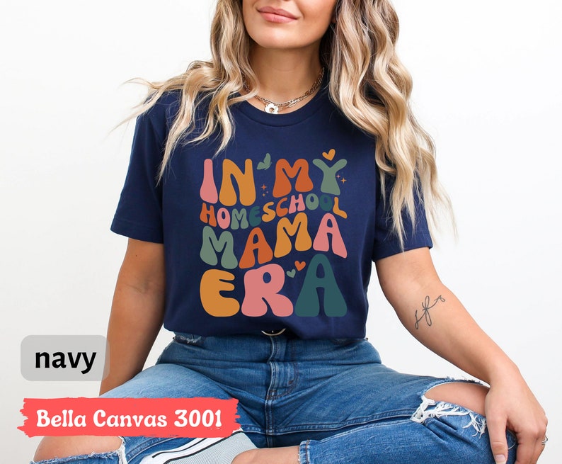 May include: A navy blue t-shirt with the text "In My Homeschool Mama Era" in colorful, wavy letters. The shirt is being worn by a person with long blonde hair.