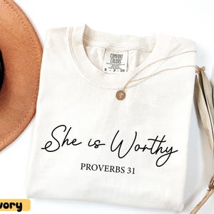 May include: Ivory-colored t-shirt with the text "She is Worthy" and "Proverbs 31" in black script. A gold necklace with a pendant rests on the shirt. A brown hat and dried plant are in the background.