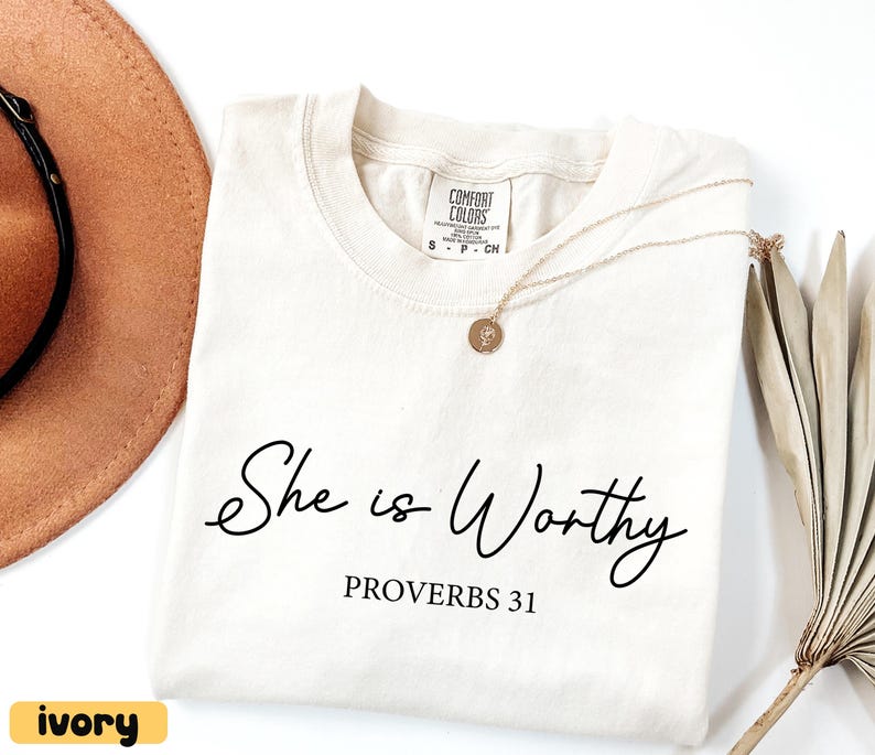 May include: Ivory-colored t-shirt with the text "She is Worthy" and "Proverbs 31" in black script. A gold necklace with a pendant rests on the shirt. A brown hat and dried plant are in the background.