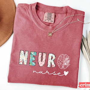 Puede incluir: Camiseta de color carmes&iacute; con la palabra "NEURO" en letras florales y rosas, y un gr&aacute;fico de cerebro con flores. La palabra "nurse" est&aacute; escrita en cursiva debajo. Un collar plateado con un colgante est&aacute; sobre la camiseta.