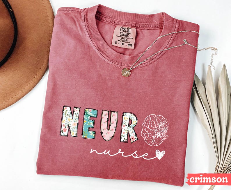 Puede incluir: Camiseta de color carmes&iacute; con la palabra "NEURO" en letras florales y rosas, y un gr&aacute;fico de cerebro con flores. La palabra "nurse" est&aacute; escrita en cursiva debajo. Un collar plateado con un colgante est&aacute; sobre la camiseta.
