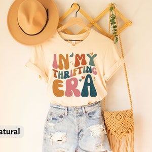 May include: A cream-colored t-shirt with the text "IN MY THRIFTING ERA" in colorful, retro-style lettering. The shirt is displayed on a wooden hanger, paired with denim shorts and a woven bag. A tan hat rests on the left.