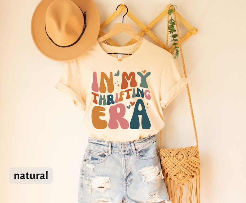 May include: A cream-colored t-shirt with the text "IN MY THRIFTING ERA" in colorful, retro-style lettering. The shirt is displayed on a wooden hanger, paired with denim shorts and a woven bag. A tan hat rests on the left.