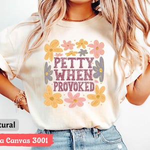 May include: A cream-colored t-shirt with the text "PETTY WHEN PROVOKED" in a purple, retro font, surrounded by colorful floral graphics in yellow, pink, and purple. The shirt is made of Bella Canvas 3001 material.