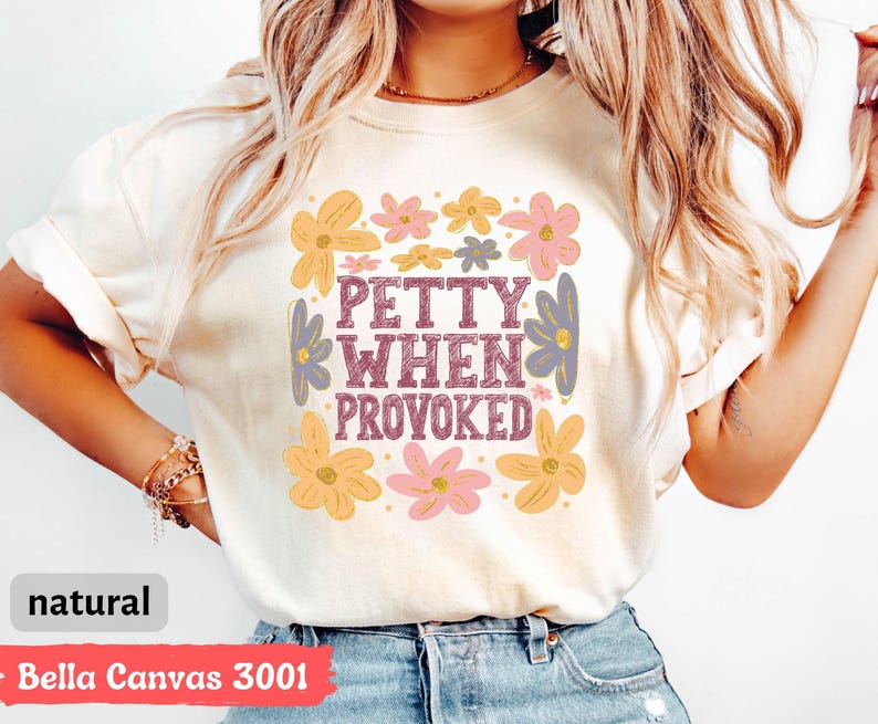 May include: A cream-colored t-shirt with the text "PETTY WHEN PROVOKED" in a purple, retro font, surrounded by colorful floral graphics in yellow, pink, and purple. The shirt is made of Bella Canvas 3001 material.