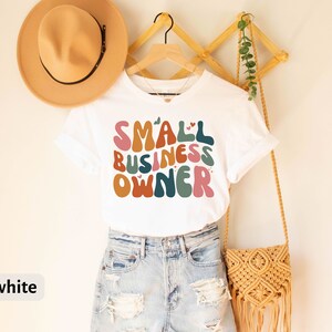 May include: White t-shirt with the text "SMALL BUSINESS OWNER" in colorful, retro-style lettering. The shirt is on a hanger and has a small heart design above the word "SMALL".