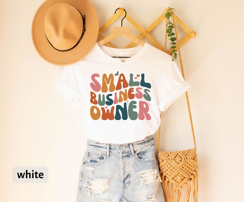 May include: White t-shirt with the text "SMALL BUSINESS OWNER" in colorful, retro-style lettering. The shirt is on a hanger and has a small heart design above the word "SMALL".
