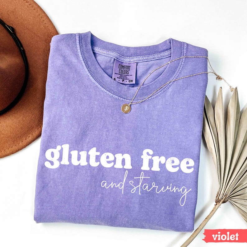 Puede incluir: Camiseta violeta Comfort Colors con las palabras "gluten free and starving" en blanco. Un collar dorado con un colgante est&aacute; sobre la camiseta. Un sombrero marr&oacute;n y una planta seca est&aacute;n en el fondo.