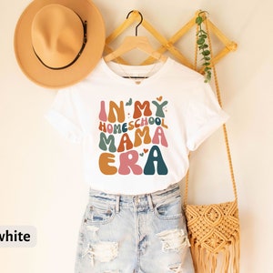 May include: White t-shirt with the text "In My Homeschool Mama Era" in colorful, retro-style lettering. The shirt is being modeled on a person wearing denim shorts.
