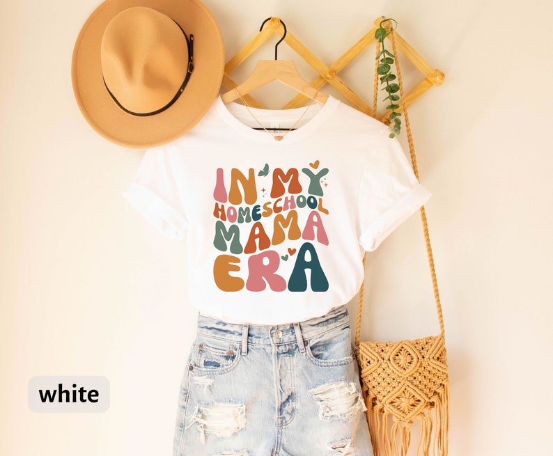 May include: White t-shirt with the text "In My Homeschool Mama Era" in colorful, retro-style lettering. The shirt is being modeled on a person wearing denim shorts.
