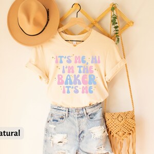 Peut inclure: Un t-shirt beige clair avec le texte "It's me, Hi I'm the Baker It's me" en lettres roses et bleues avec des accents de paillettes. Le t-shirt est suspendu &agrave; un cintre en bois avec un chapeau de paille et un sac tiss&eacute;.