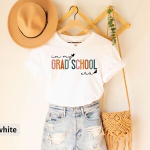 May include: White t-shirt with the text "in my GRAD SCHOOL era" in rainbow colors. The shirt is hanging on a wooden rack with a brown hat and a woven bag.