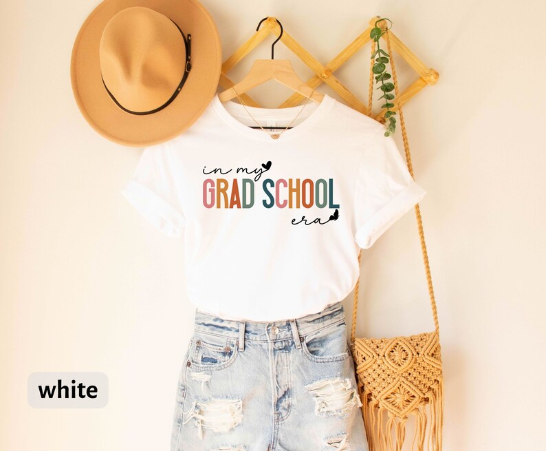 May include: White t-shirt with the text "in my GRAD SCHOOL era" in rainbow colors. The shirt is hanging on a wooden rack with a brown hat and a woven bag.