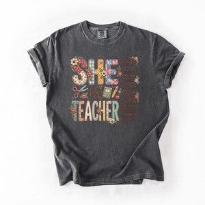 May include: Dark gray t-shirt with the words "SHE IS TEACHER" in a colorful, floral design. The design includes scissors, a book, and a pencil. Additional words like "strong" and "brave" are also included.