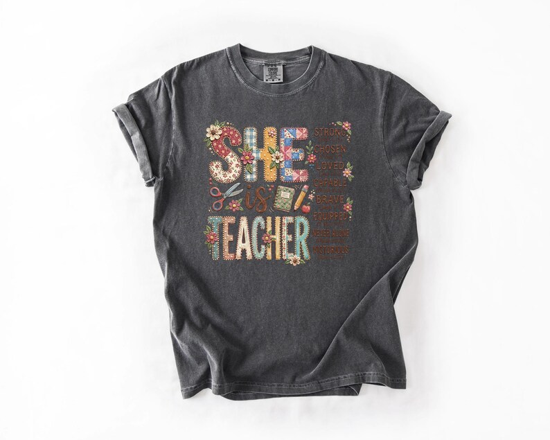 May include: Dark gray t-shirt with the words "SHE IS TEACHER" in a colorful, floral design. The design includes scissors, a book, and a pencil. Additional words like "strong" and "brave" are also included.