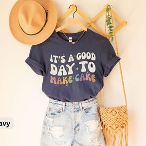 Peut inclure: T-shirt bleu marine avec le texte "It's a good day to make cake" en couleurs arc-en-ciel et des &eacute;toiles. Le t-shirt est sur un cintre et a un chapeau de paille et un sac tiss&eacute; &agrave; c&ocirc;t&eacute;.