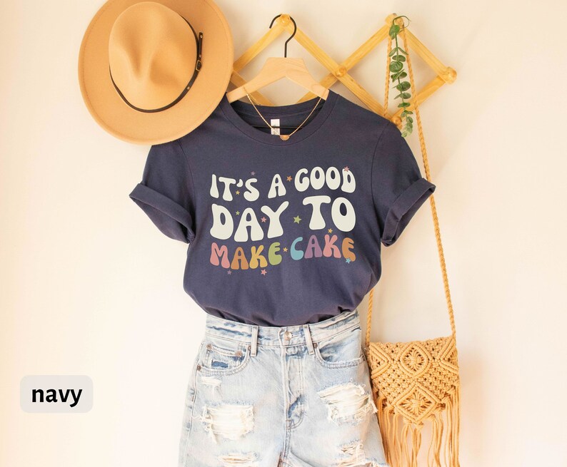 Peut inclure: T-shirt bleu marine avec le texte "It's a good day to make cake" en couleurs arc-en-ciel et des &eacute;toiles. Le t-shirt est sur un cintre et a un chapeau de paille et un sac tiss&eacute; &agrave; c&ocirc;t&eacute;.