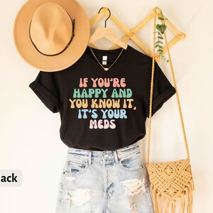 May include: Black t-shirt with the text "If you're happy and you know it, it's your meds" in rainbow colors. The shirt is being modeled with a straw hat and a denim skirt.