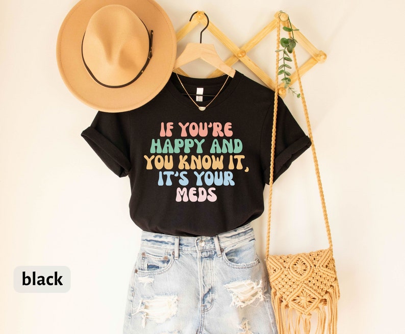 May include: Black t-shirt with the text "If you're happy and you know it, it's your meds" in rainbow colors. The shirt is being modeled with a straw hat and a denim skirt.