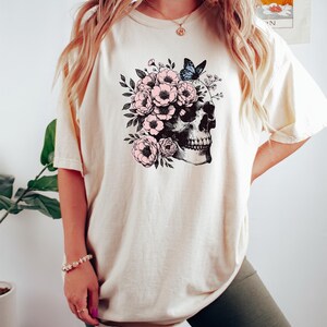 May include: A white t-shirt with a graphic of a skull with pink flowers and a blue butterfly. The skull is surrounded by flowers and leaves.