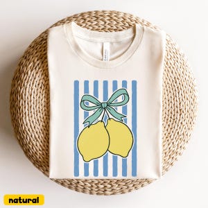 May include: A white t-shirt with a blue and white striped design featuring two yellow lemons tied together with a blue bow.