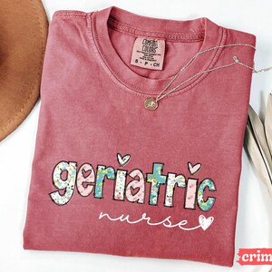 Pode incluir: Camiseta cor carmesim com as palavras "geriatric nurse" em um design floral e de cora&ccedil;&atilde;o. Um colar de prata com um pingente est&aacute; sobre a camiseta. A camiseta tem uma etiqueta Comfort Colors.