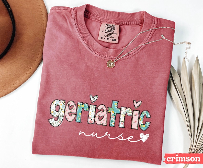 Pode incluir: Camiseta cor carmesim com as palavras "geriatric nurse" em um design floral e de cora&ccedil;&atilde;o. Um colar de prata com um pingente est&aacute; sobre a camiseta. A camiseta tem uma etiqueta Comfort Colors.