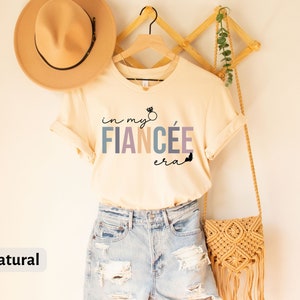 May include: A beige t-shirt with the text "in my fianc&eacute;e era" in colorful letters with a small ring and butterfly graphic. The shirt is hanging on a wooden hanger with a straw bag and a hat.