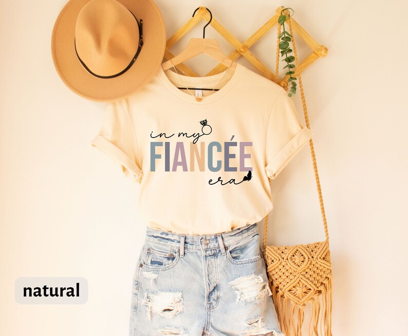 May include: A beige t-shirt with the text "in my fianc&eacute;e era" in colorful letters with a small ring and butterfly graphic. The shirt is hanging on a wooden hanger with a straw bag and a hat.