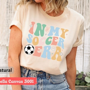 Może przedstawiać: Beżowa koszulka z napisem "In My Soccer Era" w kolorowych, retro literach. W projekcie znajduje się piłka nożna.
