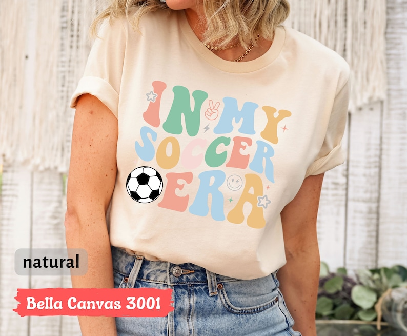 Może przedstawiać: Beżowa koszulka z napisem "In My Soccer Era" w kolorowych, retro literach. W projekcie znajduje się piłka nożna.