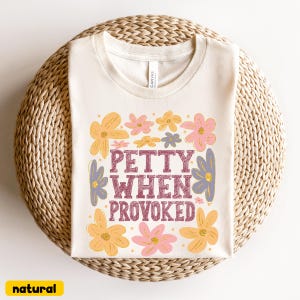 May include: Cream-colored t-shirt with the text "PETTY WHEN PROVOKED" in maroon, surrounded by yellow, pink, and purple floral graphics. The shirt is displayed on a woven, circular surface. The word "natural" is in a yellow box.