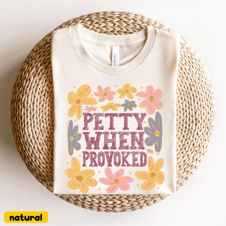 May include: Cream-colored t-shirt with the text "PETTY WHEN PROVOKED" in maroon, surrounded by yellow, pink, and purple floral graphics. The shirt is displayed on a woven, circular surface. The word "natural" is in a yellow box.