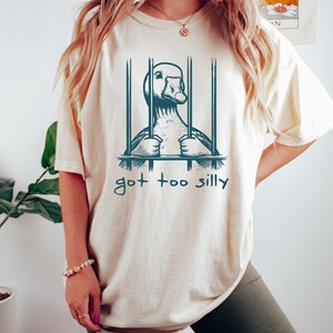 May include: A white t-shirt with a graphic of a goose in a cage. The text "got too silly" is printed below the graphic.