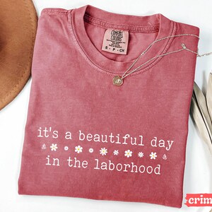 May include: Crimson-colored t-shirt with the phrase "it's a beautiful day in the laborhood" in white script. The shirt features small flower graphics. A silver necklace with a pendant is draped across the shirt. A brown hat is visible in the upper left corner.