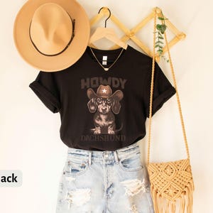 Puede incluir: Camiseta negra con un dise&ntilde;o de un perro salchicha con sombrero de vaquero y el texto "HOWDY DACHSHUND". La camiseta se combina con pantalones cortos vaqueros azules claros, un sombrero beige y un bolso de macram&eacute;.