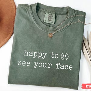 May include: A green t-shirt with the text "happy to see your face" and a smiley face graphic printed in white.