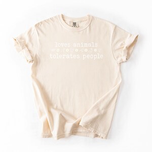 May include: A beige t-shirt with the text "loves animals tolerates people" printed on the front. The text is surrounded by small flower and star graphics.