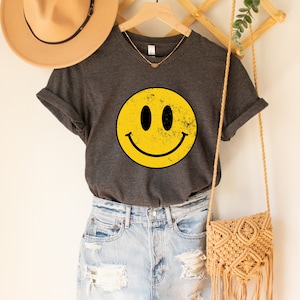 May include: A dark grey t-shirt with a yellow smiley face graphic. The smiley face has a distressed look and is centred on the shirt. The shirt is hanging on a wooden hanger.