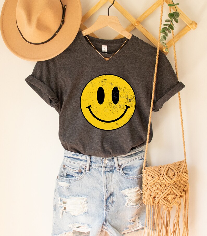 May include: A dark grey t-shirt with a yellow smiley face graphic. The smiley face has a distressed look and is centred on the shirt. The shirt is hanging on a wooden hanger.