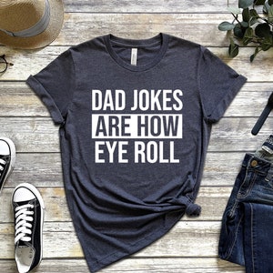 May include: A dark gray t-shirt with white text that reads "Dad Jokes Are How Eye Roll".