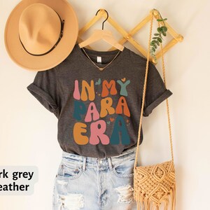 May include: Dark grey heather t-shirt with the text "In My Para Era" in colorful, retro-style lettering. The shirt is hanging on a wooden hanger with a straw bag and a hat.