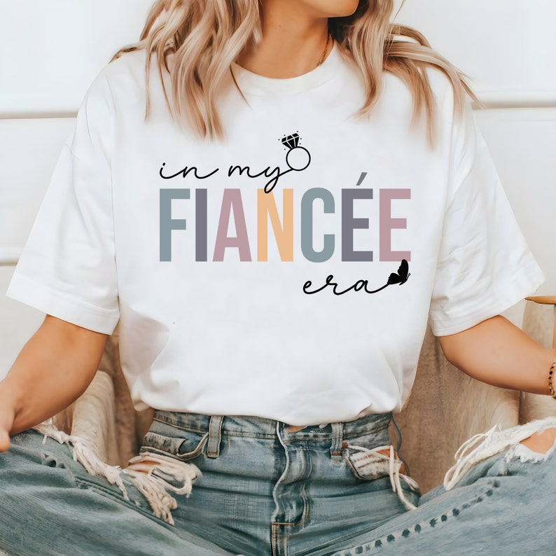 May include: White t-shirt with the text "in my fianc&eacute;e era" in rainbow colors with a diamond ring graphic and a butterfly.